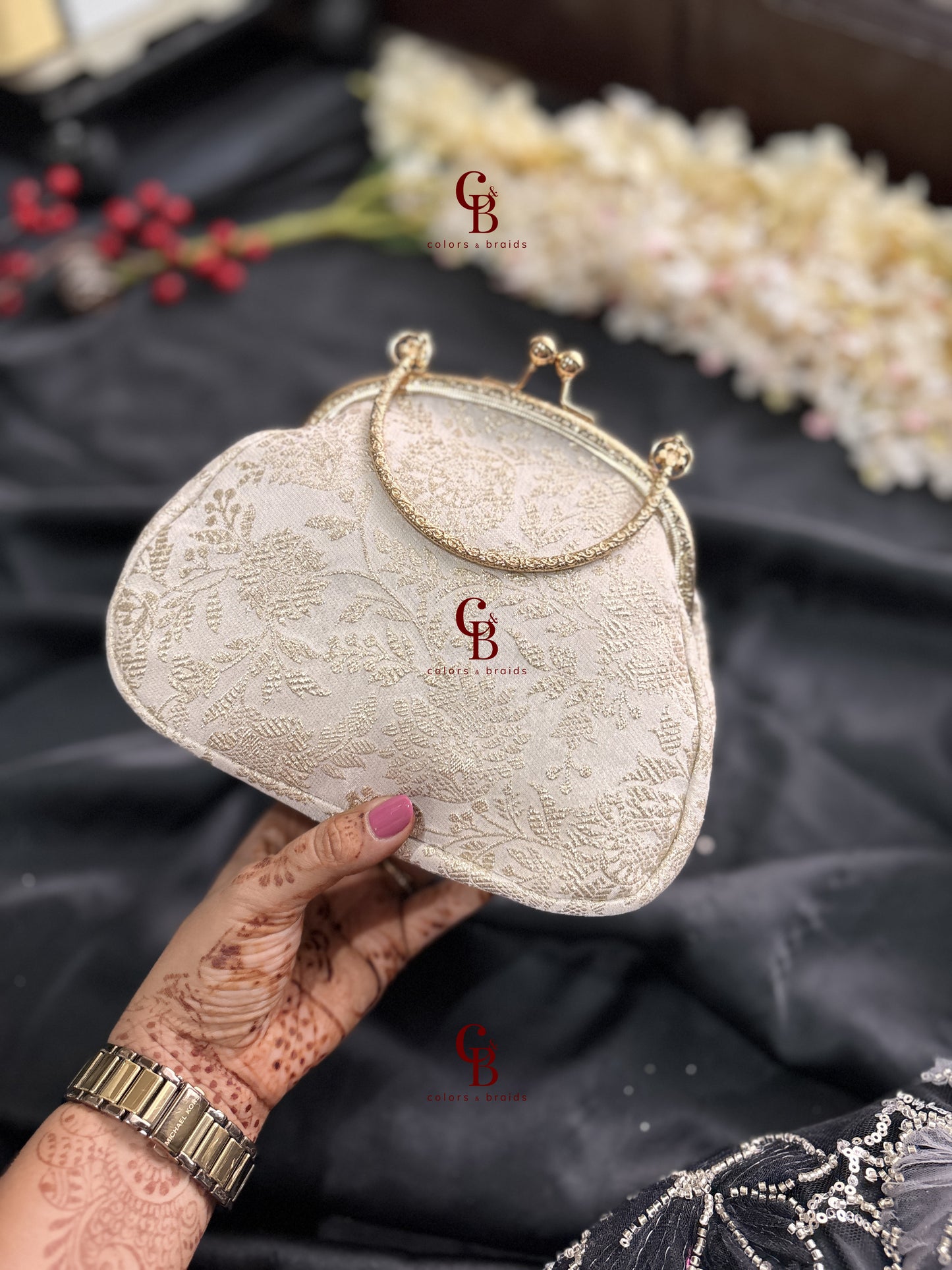 White embroidered clutch bag held by a hand with henna designs on a black fabric background.